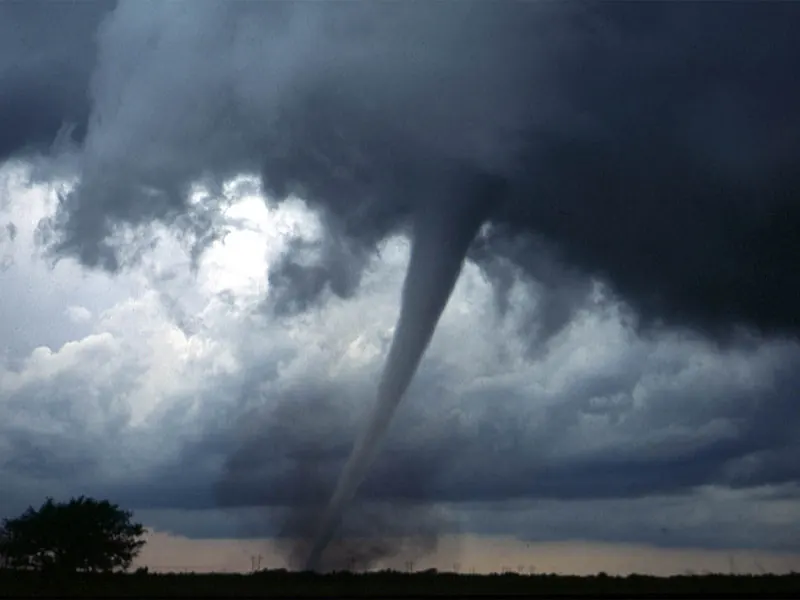 A long, thin, grey tornado reaching down from the storm to the ground across open country