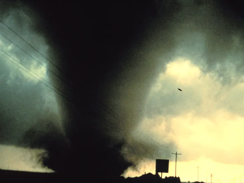 A wide, dark tornado touching the ground under a stormy sky, with dust swirling at its base