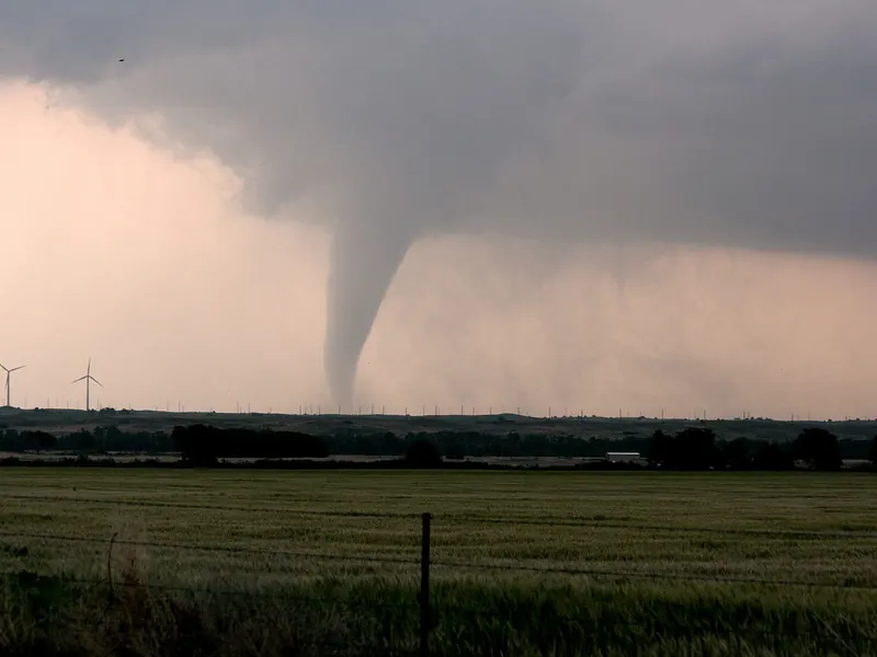 A thin grey tornado funnel reaching down from a dark cloud base to flat open farmland