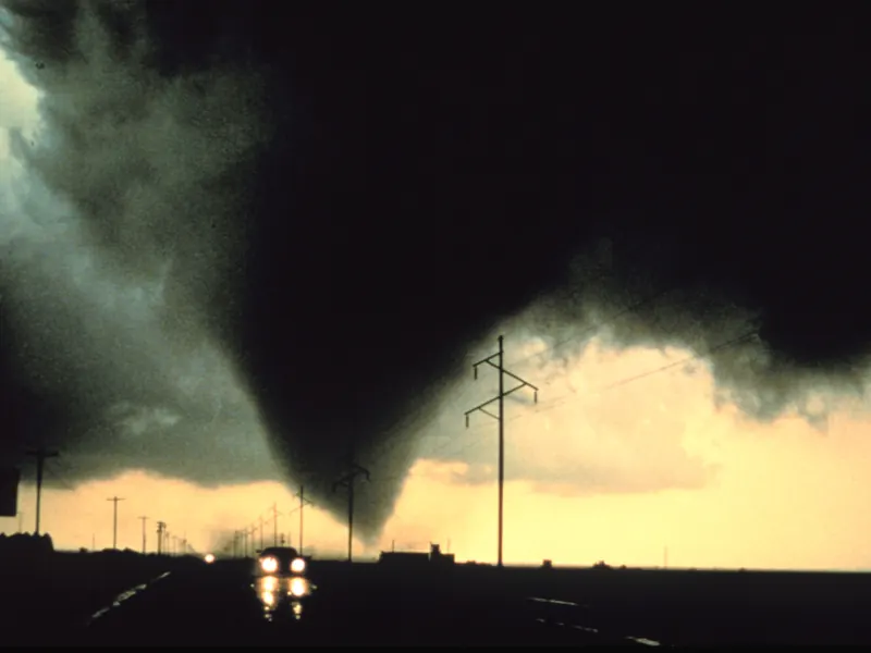 A dark tornado funnel against an orange evening sky, with power poles and a road in silhouette