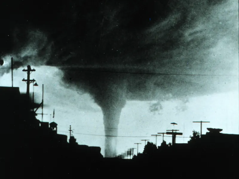 An old black-and-white photo of a tornado approaching a small town, with buildings and poles in silhouette