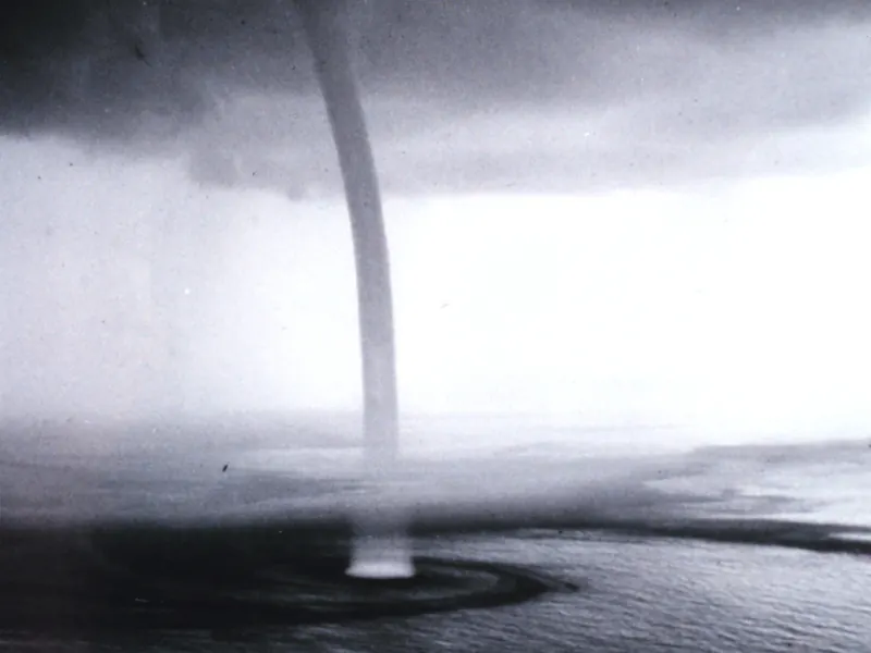 A thin, pale column of spray rising from a dark sea to the cloud above, with a ring of spray at its base